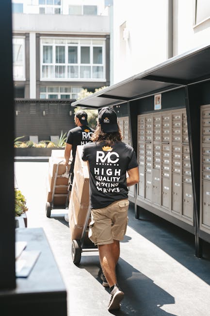 Two removal service workers from Removals Richmond upon Thames are outside a residential building, loading cardboard boxes and large parcels onto a trolley. The workers are wearing black caps and blac