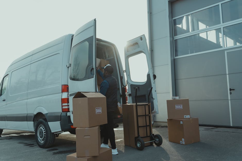 Two removal service workers from Removals Richmond upon Thames are outside a residential building, loading cardboard boxes and large parcels onto a trolley. The workers are wearing black caps and blac